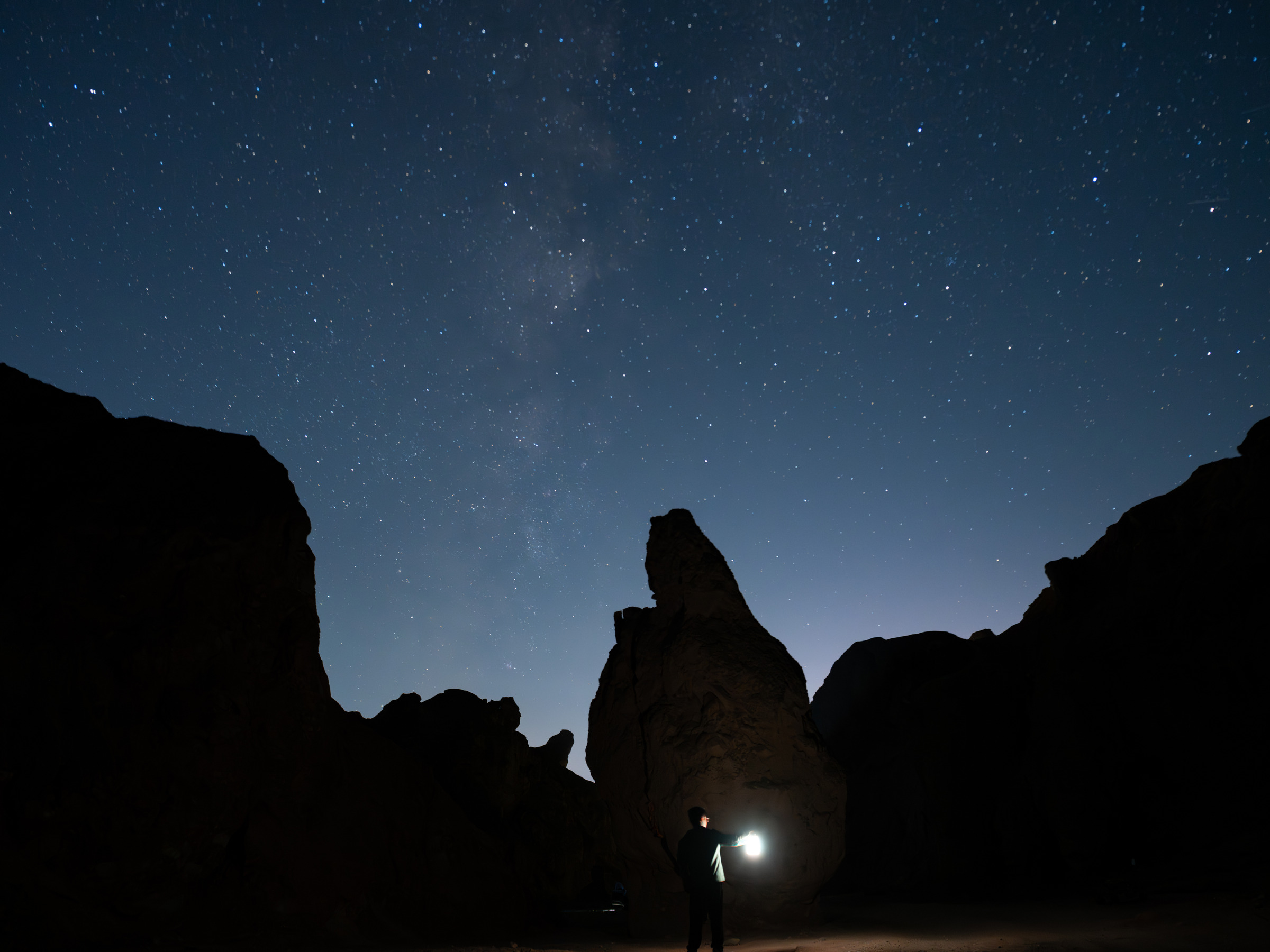 黑色岩壁围出夜空，银河横过上方，谷中人物举着暖色灯光。