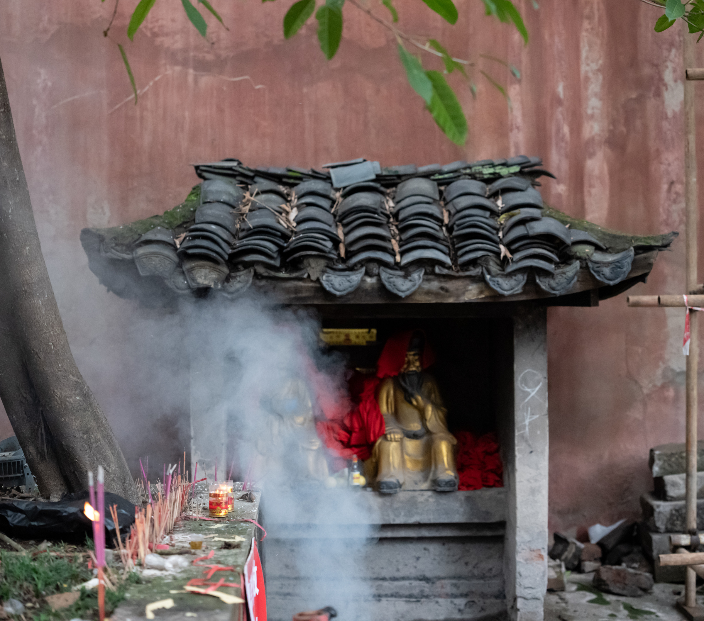神龛与香炉前升起薄烟，红墙、金色细节和绿叶交织。